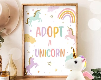 May include: A framed poster with a white background and colorful illustrations of unicorns, rainbows, and stars. The poster says "Adopt a Unicorn". A white plush unicorn toy with a rainbow mane and tail is sitting in front of the poster.
