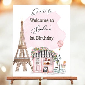 May include: A pink and white welcome sign with a watercolor background. The sign says "Oh la la Welcome to Sophie's 1st Birthday". The sign also features a drawing of the Eiffel Tower, a bakery, and a bicycle.