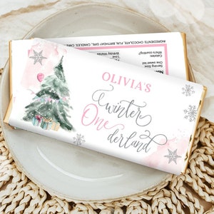 May include: A chocolate bar wrapper with a winter theme. The wrapper features a watercolor illustration of a Christmas tree, gifts, and snowflakes. The text "Olivia's Winter One derland" is displayed in pink and gray script.