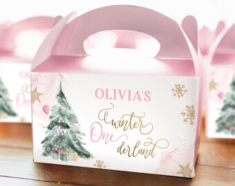 May include: Pink and gold favor boxes with a winter wonderland theme. The boxes feature a watercolor illustration of a Christmas tree with presents and a balloon. The text on the box reads "Olivia's Winter Wonderland".