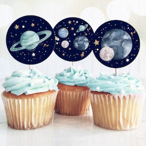 May include: Three blue and white cupcake toppers with a space theme. The toppers feature planets, stars, and a rocket ship on a dark blue background.