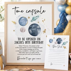 Op de afbeelding: Een ingelijst bord met de titel Time capsule met aquarelillustraties van planeten en een raket. De tekst luidt To be opened on Jacob's 18th birthday, gevolgd door een gedicht en een verzoek om te ondertekenen. Een bijpassende kaart staat ervoor.