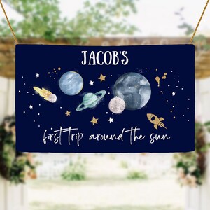 Puede incluir: Una pancarta azul marino con estrellas doradas y planetas, un cohete y el texto "JACOB'S first trip around the sun".
