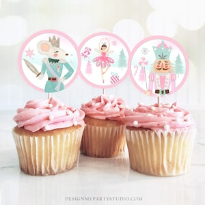 May include: Three pink frosted cupcakes with white sprinkles topped with round, pink and white paper cupcake toppers featuring illustrations of a mouse soldier, a ballerina, and a nutcracker.