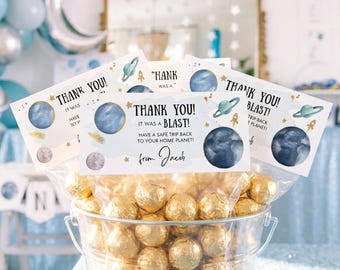 May include: A clear plastic bucket filled with round gold foil-wrapped candies, topped with a white rectangular card featuring space-themed illustrations of planets, stars, and rockets, with the text: THANK YOU! IT WAS A BLAST! FROM JACOB.