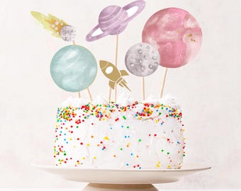 Puede incluir: Un pastel con glaseado blanco cubierto de chispas de colores y adornos de papel con temática espacial, incluyendo un cohete dorado, un planeta azul claro, un planeta morado con anillos, una luna gris, un planeta rosa y un cometa amarillo.