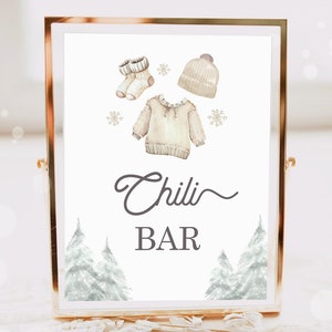 May include: A white sign with a gold frame, featuring a watercolor illustration of a sweater, hat, and socks. The sign reads "Chili Bar" in a gray script font, with a decorative flourish under the word "Chili". The background is white with a few snowflakes scattered around the illustration.