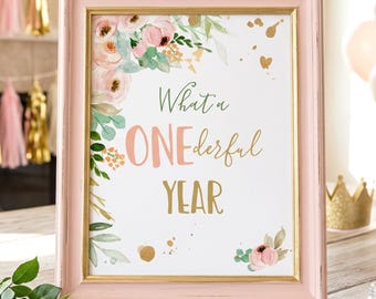 May include: A light pink wooden frame containing a white sign with watercolor floral accents in pink and green, gold paint splatters, and the text "What a ONEderful YEAR" written in a mix of green, pink, and gold script and block lettering.