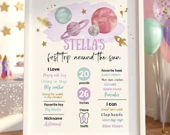 May include: A white framed sign titled Stella's first trip around the sun, featuring watercolor planets, a rocket, and text detailing milestones like 20 pounds, 26 inches, and 6 teeth, alongside lists of favorite foods, toys, and activities.