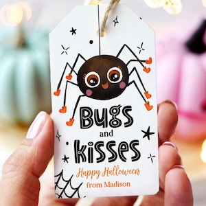 May include: White Halloween gift tag with a cartoon spider illustration. The tag reads "Bugs and Kisses" and "Happy Halloween from Madison". The spider is brown with orange accents. The tag is held by a person.