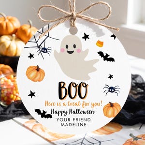 May include: A round white hanging ornament featuring a central beige ghost illustration, orange pumpkins, black spiders, bats, stars, and candy corn. Text reads: BOO, Here is a treat for you!, Happy Halloween, YOUR FRIEND MADELINE. Includes twine.
