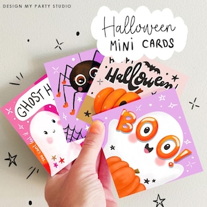 May include: A hand holds a stack of Halloween mini cards. Each card features a different spooky illustration, including a ghost, spider, and pumpkin. The cards are in shades of pink, purple, and orange, with the words "Halloween" and "BOO" written on them.
