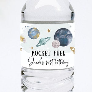 May include: Water bottle label with a space theme. The label features planets, stars, and a rocket ship. The text on the label reads "Rocket Fuel Jacob's first birthday".