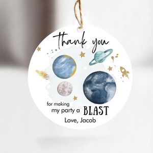 May include: Round thank you tag with a space theme. It features planets, stars, and a rocket ship. The text reads "Thank you for making my party a BLAST Love, Jacob".