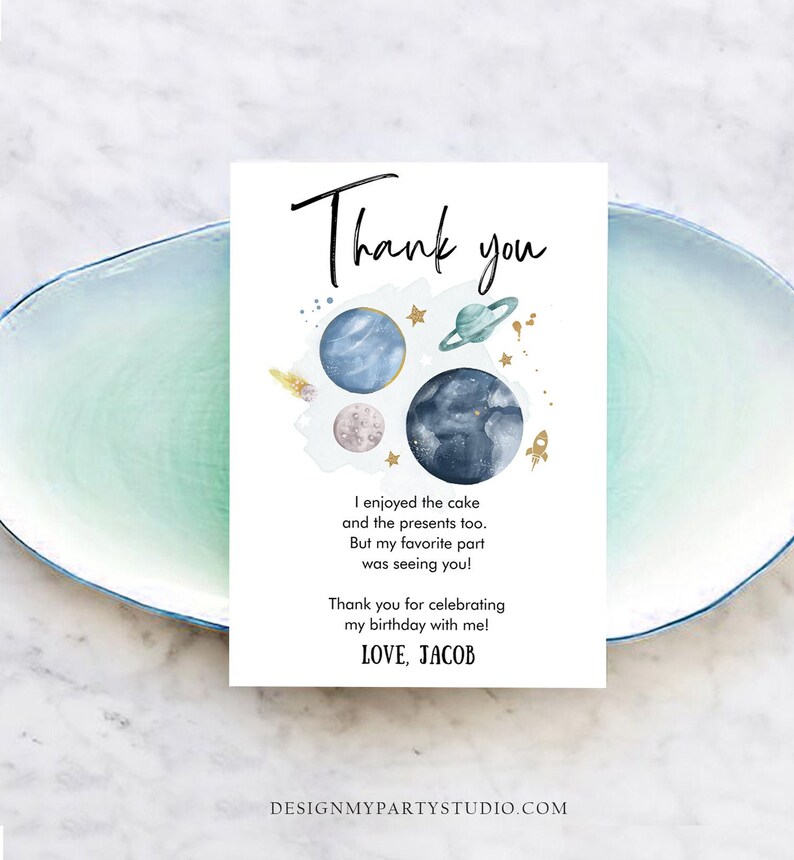 May include: A white thank you card with a watercolor space theme. The card features planets, stars, and a rocket ship illustration. Text includes "Thank you" and a personalized message. The card is on a light blue plate.