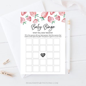 May include: A white Baby Bingo game card with a strawberry design. The card has a grid of squares, a heart in the center, and the text "Baby Bingo" and "What will Emily receive?" at the top. A pencil and envelope are in the background.
