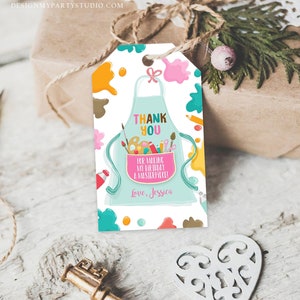 May include: A gift tag featuring a light blue apron design, paint splatters, and the text "Thank You for Making My Birthday a Masterpiece!" The tag includes paintbrushes, pencils, and a paint palette. The tag is on a gift.