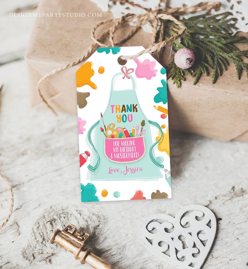 May include: A gift tag featuring a light blue apron design, paint splatters, and the text "Thank You for Making My Birthday a Masterpiece!" The tag includes paintbrushes, pencils, and a paint palette. The tag is on a gift.