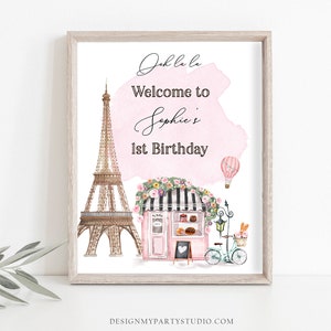 以下が含まれることがあります： ピンクと白の水彩画の背景に、「Oh la la ソフィーの1歳の誕生日へようこそ」というテキストが書かれています。画像には、エッフェル塔、花輪で飾られたピンクのベーカリー、熱気球、パンの入ったバスケットが付いた自転車の漫画イラストも描かれています。