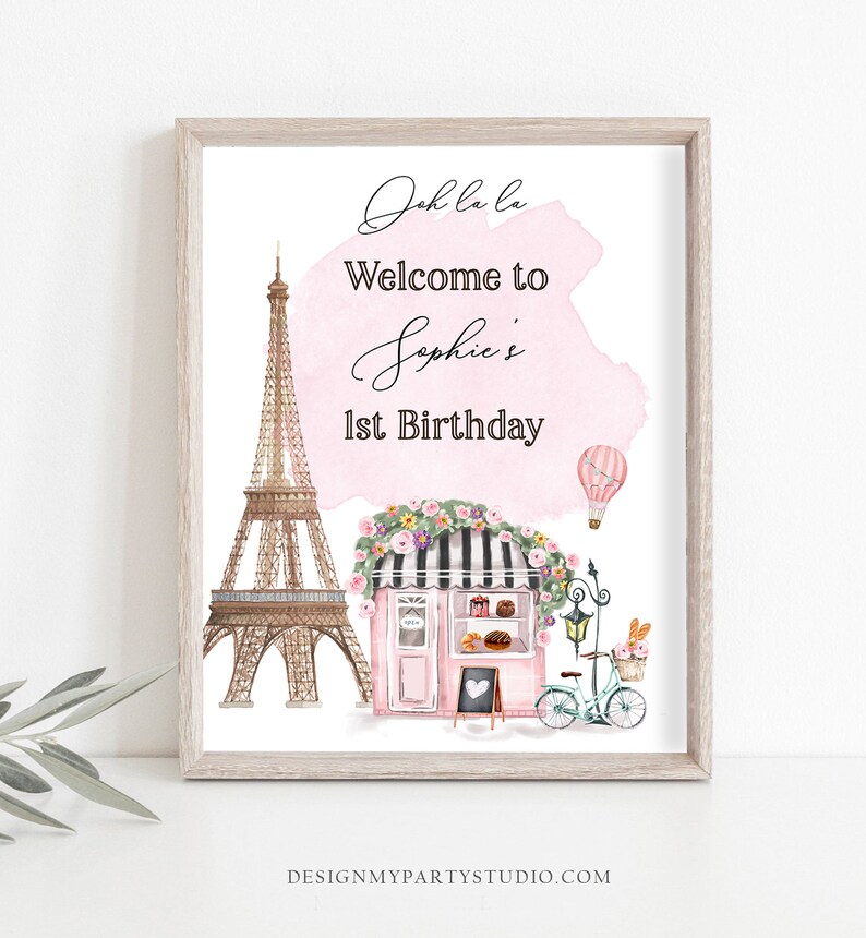 以下が含まれることがあります： ピンクと白の水彩画の背景に、「Oh la la ソフィーの1歳の誕生日へようこそ」というテキストが書かれています。画像には、エッフェル塔、花輪で飾られたピンクのベーカリー、熱気球、パンの入ったバスケットが付いた自転車の漫画イラストも描かれています。