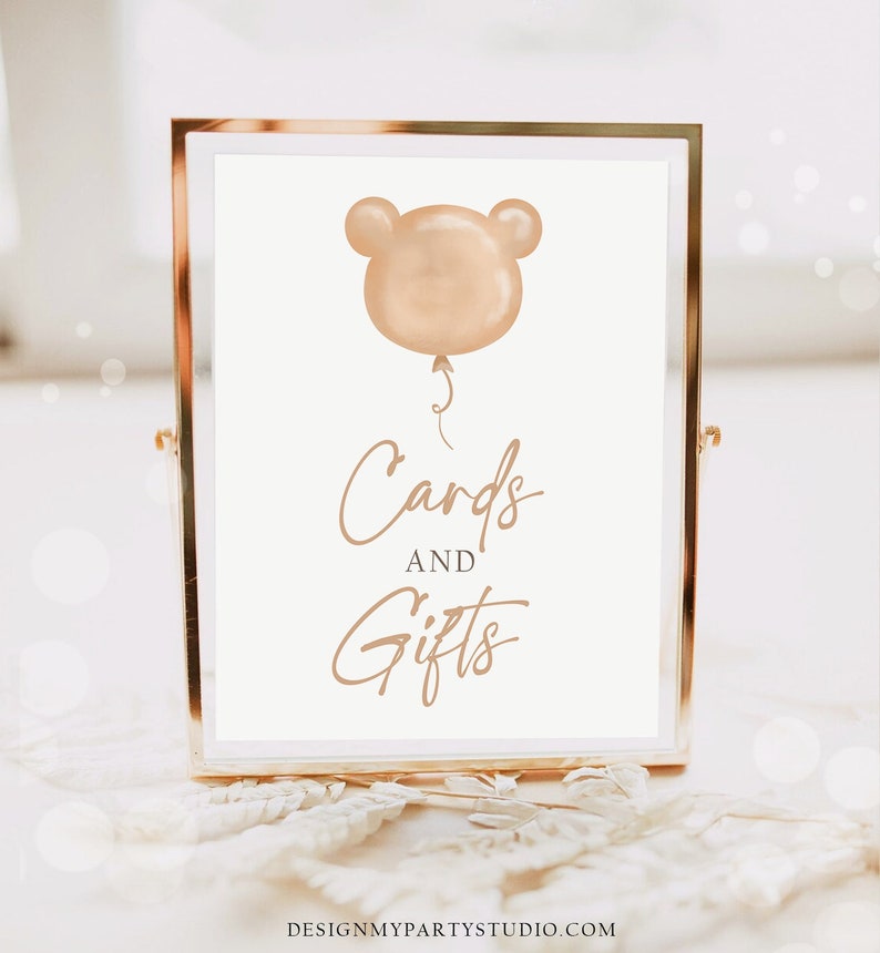 May include: A gold-framed sign with a white background and a brown balloon with ears. The sign reads "Cards and Gifts".