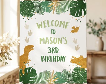 Op de afbeelding: Een rechthoekig bord met de tekst WELCOME TO MASON'S 3RD BIRTHDAY in groene letters. Het ontwerp bevat illustraties van twee gele dinosaurussen, beige dinosaurusvoetafdrukken en groene tropische palmbladeren op een witte achtergrond.