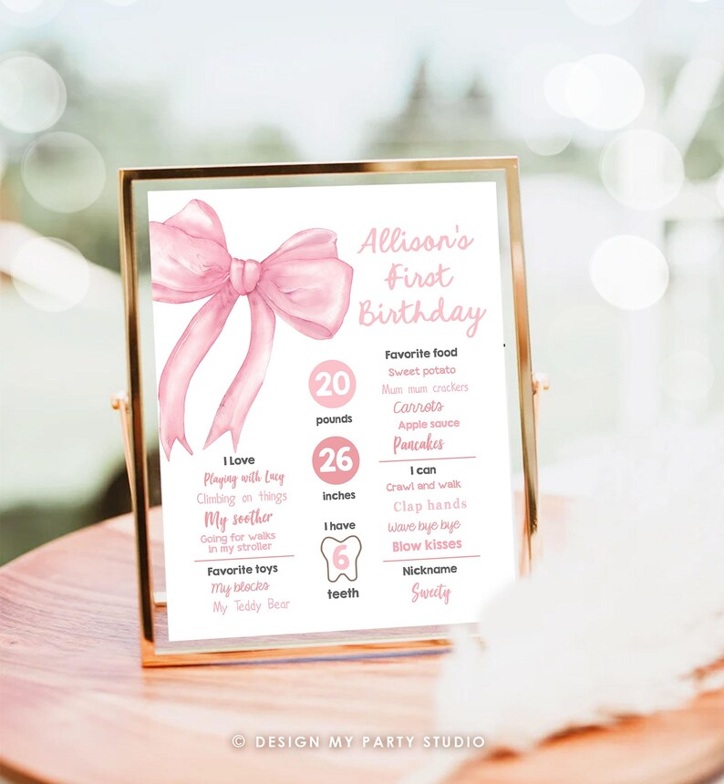 May include: Pink and white first birthday milestone poster with a large pink bow at the top. The poster lists the child's age, weight, height, and other milestones. The text on the poster reads "Allison's First Birthday".