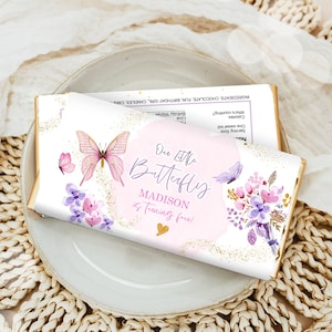 May include: A chocolate bar with a custom wrapper featuring a butterfly theme. The wrapper has a pink and gold design with the text "Our Little Butterfly" and "Madison is turning four!" on a white background. The bar rests on a gray plate.
