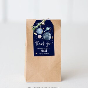 May include: A blue and gold thank you gift tag with a space theme. The tag features planets, stars, and a rocket ship. The text on the tag reads "Thank you for making my party a BLAST. Love, Jackson."