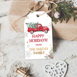 Puede incluir: Una etiqueta de regalo blanca con un cami&oacute;n rojo que transporta un &aacute;rbol de Navidad. La etiqueta dice "Happy Holidays!" y "From The Smith's Family" en letras rojas y doradas. Copos de nieve y vegetaci&oacute;n decoran los bordes.