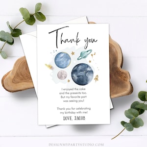 May include: A white thank you card with a space theme. The card features watercolor planets, stars, and a rocket ship. The text reads "Thank you" and a personalized message. The card is on a wooden surface with greenery.
