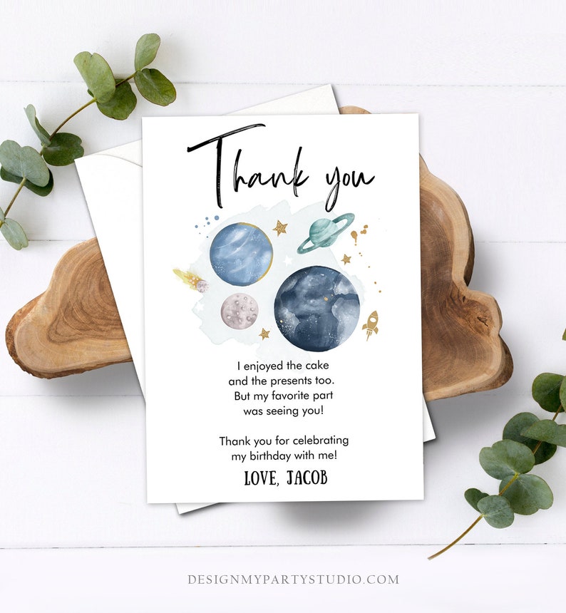 May include: A white thank you card with a space theme. The card features watercolor planets, stars, and a rocket ship. The text reads "Thank you" and a personalized message. The card is on a wooden surface with greenery.