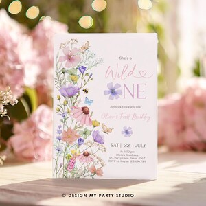 May include: A floral-themed first birthday invitation with a watercolor design. The invitation features a pink and purple floral arrangement with butterflies and bees. The text reads "She's a Wild One" and includes details for the party, including the date, time, and location. The invitation is designed by "Design My Party Studio."
