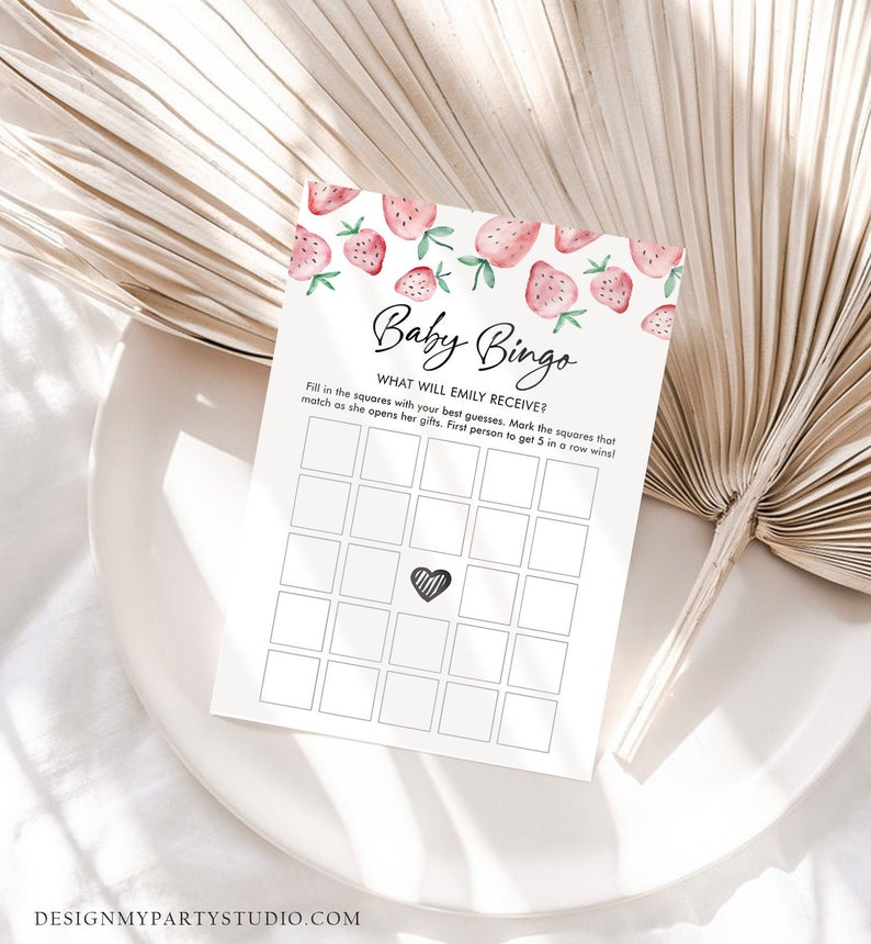 May include: A white Baby Bingo game card with a strawberry design at the top. The card has a grid of squares and the text "Baby Bingo" and "What will Emily receive?" is printed on the card. The card is on a white plate.