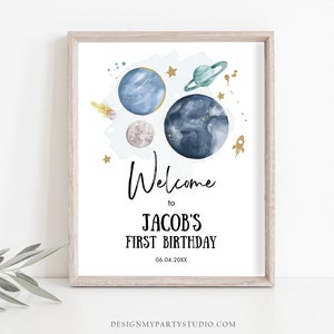 May include: A printable welcome sign for a first birthday party with a space theme. The sign features a watercolor illustration of planets, stars, and a rocket ship. The text reads "Welcome to Jacob's First Birthday 06.04.20XX".