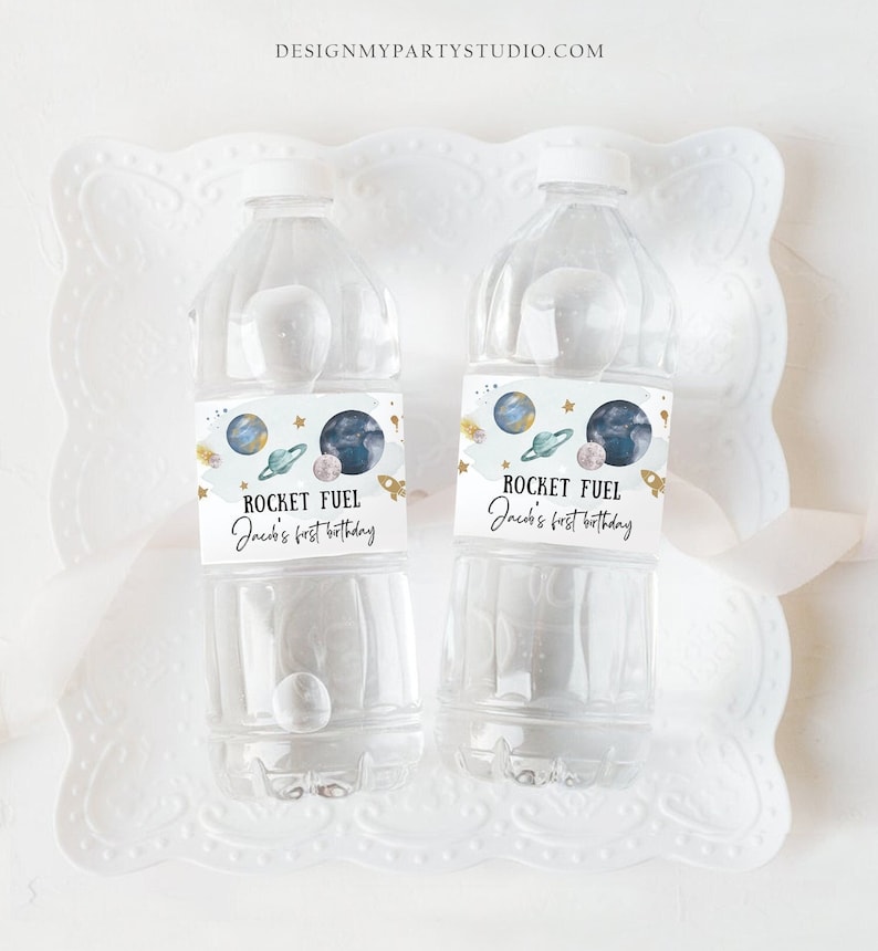 May include: Two clear plastic water bottles with blue and gold labels that say "Rocket Fuel" and "Jack's first birthday". The labels feature a watercolor illustration of planets and stars.