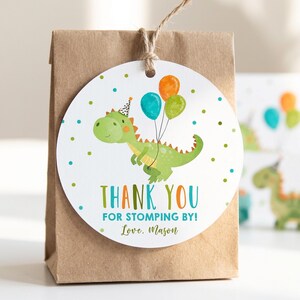 May include: A round white paper gift tag attached to a brown paper bag with twine. The tag features a green dinosaur wearing a party hat, holding orange, blue, and green balloons, with the text THANK YOU FOR STOMPING BY! Love, Mason.