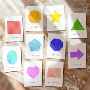 Pode incluir: Um conjunto de cartões com formas geométricas em aquarela, incluindo um quadrado vermelho, um círculo amarelo e um triângulo verde. Cada cartão apresenta o nome da forma em uma fonte clara. Os cartões estão dispostos sobre uma superfície de madeira. O texto "DESIGN MY PARTY STUDIO" é visível.