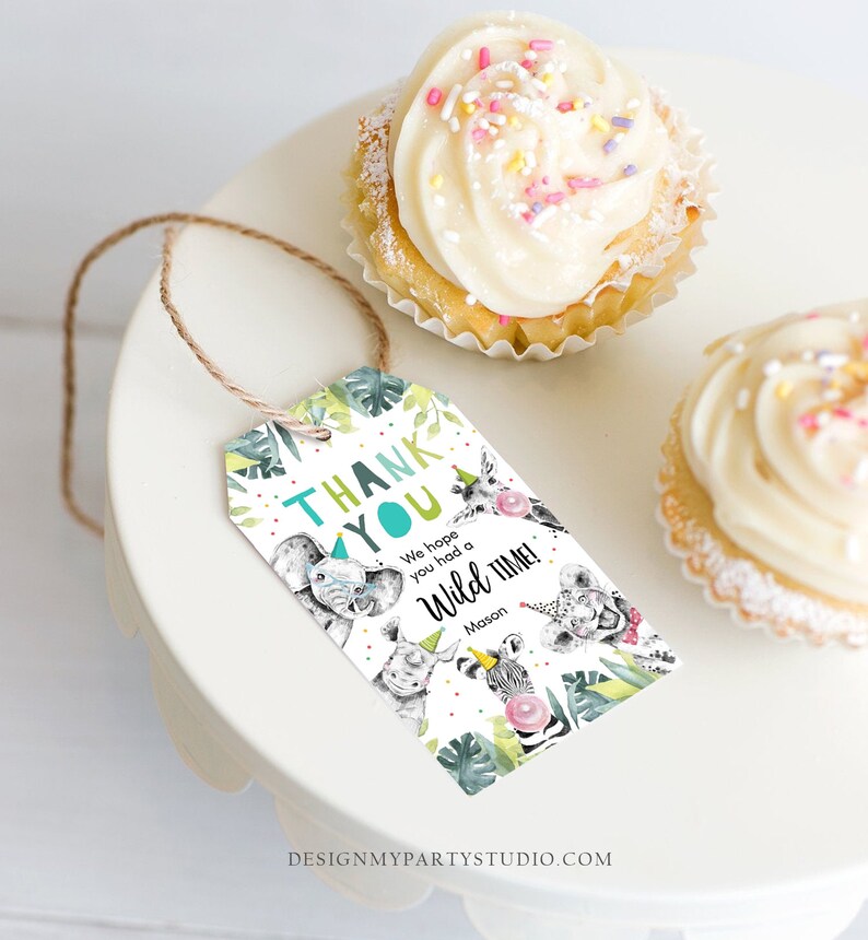 May include: A party favor tag with a jungle animal theme. The tag says "Thank You" and "We hope you had a Wild Time!" with illustrations of an elephant, rhino, zebra, giraffe, and lion. The tag is attached to a white cake stand with two cupcakes.