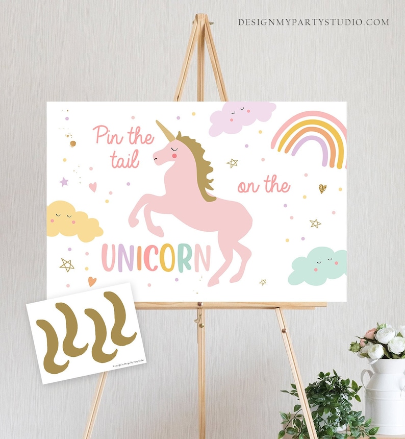 Pode incluir: Um tabuleiro de jogo branco com um unic&oacute;rnio rosa, um arco-&iacute;ris e nuvens. O texto no tabuleiro diz "Pin the tail on the unicorn". H&aacute; tr&ecirc;s caudas de unic&oacute;rnio douradas em uma folha de papel branca separada.