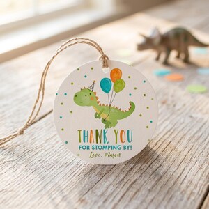 May include: A round white paper gift tag with a jute twine string featuring a green dinosaur illustration wearing a party hat and holding three balloons. The text reads THANK YOU FOR STOMPING BY! and Love, Mason, surrounded by small green and blue dots.