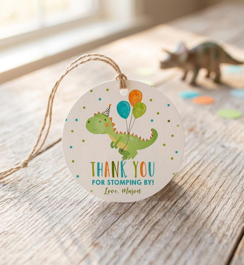 May include: A round white paper gift tag with a jute twine string featuring a green dinosaur illustration wearing a party hat and holding three balloons. The text reads THANK YOU FOR STOMPING BY! and Love, Mason, surrounded by small green and blue dots.