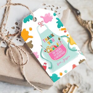 May include: A gift tag with a light blue apron design, featuring the words "Thank You" and "For making my birthday a masterpiece!" with the name "Jessica". The tag is decorated with paint splatters and art supplies.