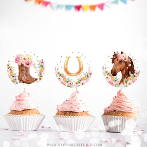 May include: Three pink cupcakes with white wrappers topped with pink frosting. Each cupcake has a round, white paper topper with a different design: a pair of brown cowboy boots with pink flowers, a gold horseshoe with pink flowers, and a brown horse with a pink flower crown.