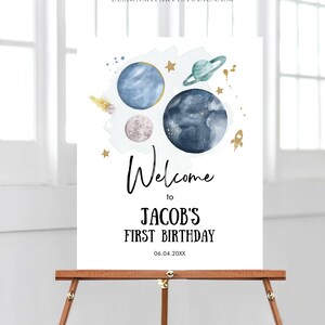 May include: Welcome to Jacob's First Birthday sign with a watercolor illustration of planets, stars, and a rocket ship. The sign is on a wooden easel.