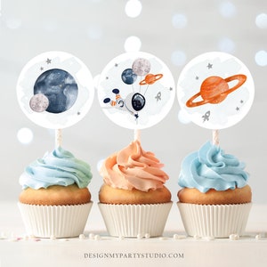 May include: Three cupcakes with pastel frosting and space-themed toppers. The toppers feature watercolor illustrations of planets, an astronaut with balloons, and rockets. The cupcakes are in white paper cups, with the text "DESIGNMYPARTYSTUDIO.COM" visible at the bottom.