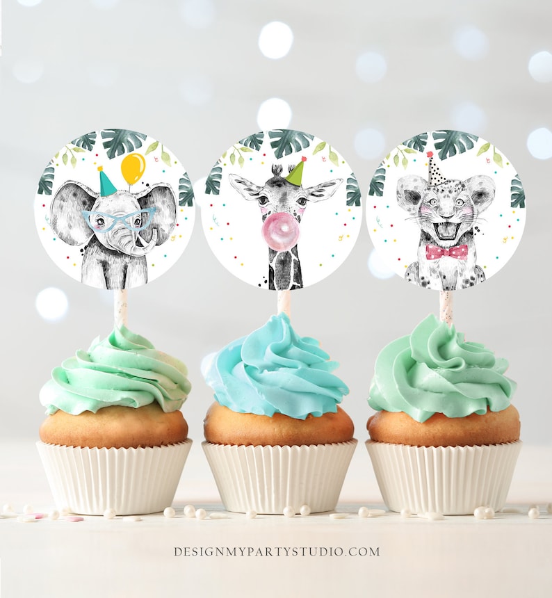 May include: Three cupcakes with blue frosting topped with round paper cupcake toppers featuring an elephant, giraffe, and lion wearing party hats. The toppers have a jungle theme with green leaves and white polka dots.