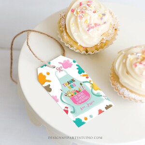 May include: A white gift tag featuring a teal apron design, paint splatters, and the words "THANK YOU." The tag includes the message "For making my birthday a masterpiece!" and the name "Jessica." Two cupcakes with white icing and sprinkles are also visible.