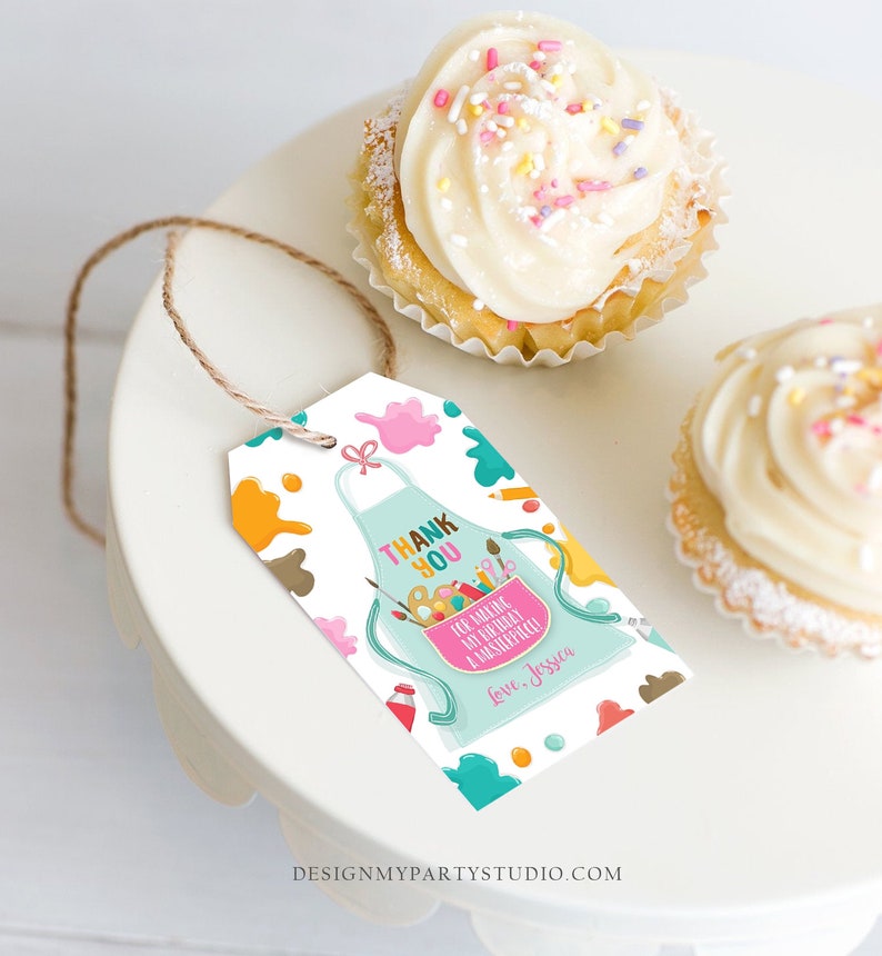May include: A white gift tag featuring a teal apron design, paint splatters, and the words "THANK YOU." The tag includes the message "For making my birthday a masterpiece!" and the name "Jessica." Two cupcakes with white icing and sprinkles are also visible.
