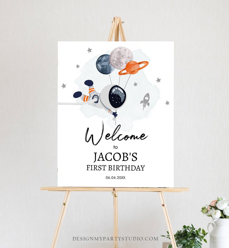May include: Welcome to Jacob's First Birthday sign with a cartoon astronaut holding balloons shaped like planets and a rocket ship. The sign is on a wooden easel.